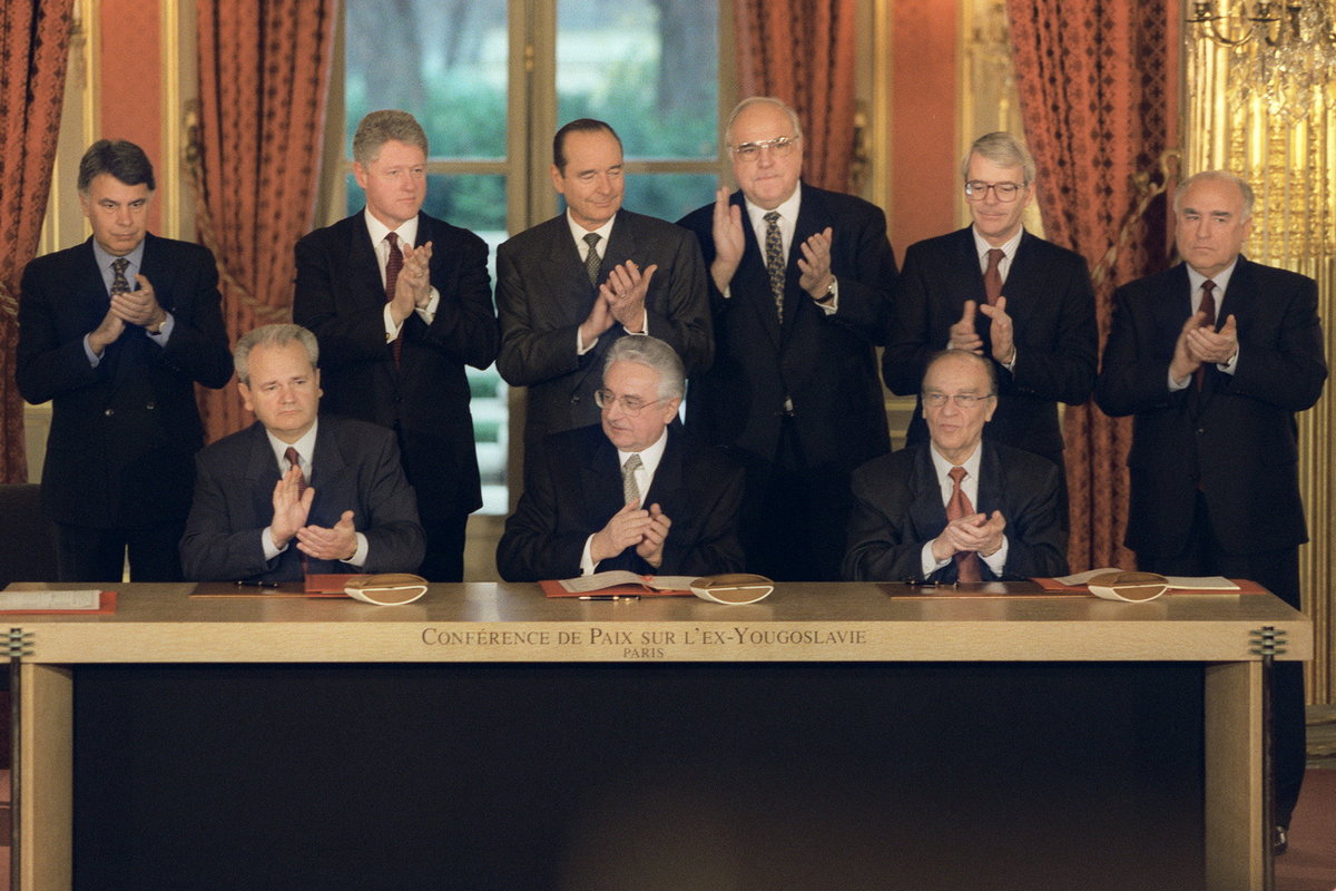 Tre menn i dress sitter ved et bord og klapper, bak står seks til dresskledde menn og klapper.