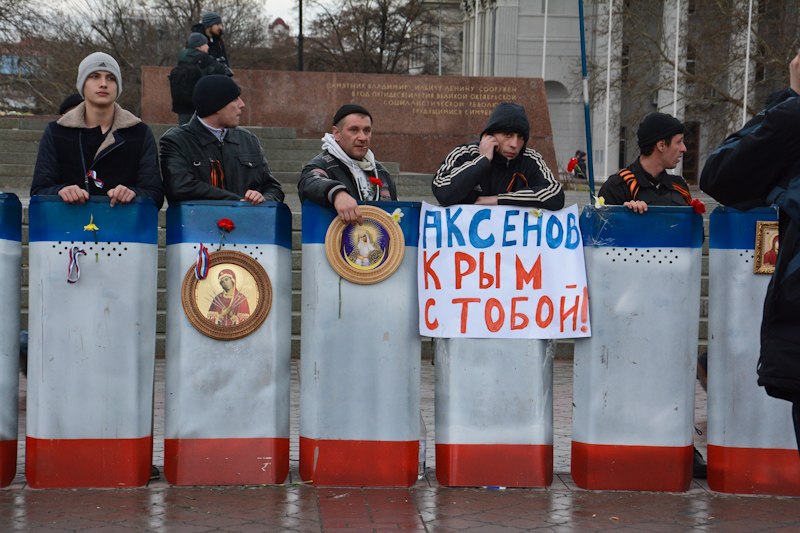 Fem personer står bak skjold malt i det pro-russiske Krim-flagget. Det har en plakat, der det står noe russisk, og noen bilder av det som fremstår som en gudelignende skikkelise 