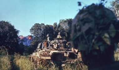 Amerikanske soldater sitter på toppen av en tanks, som kjører over et åpent gresskledd område mellom trærne.