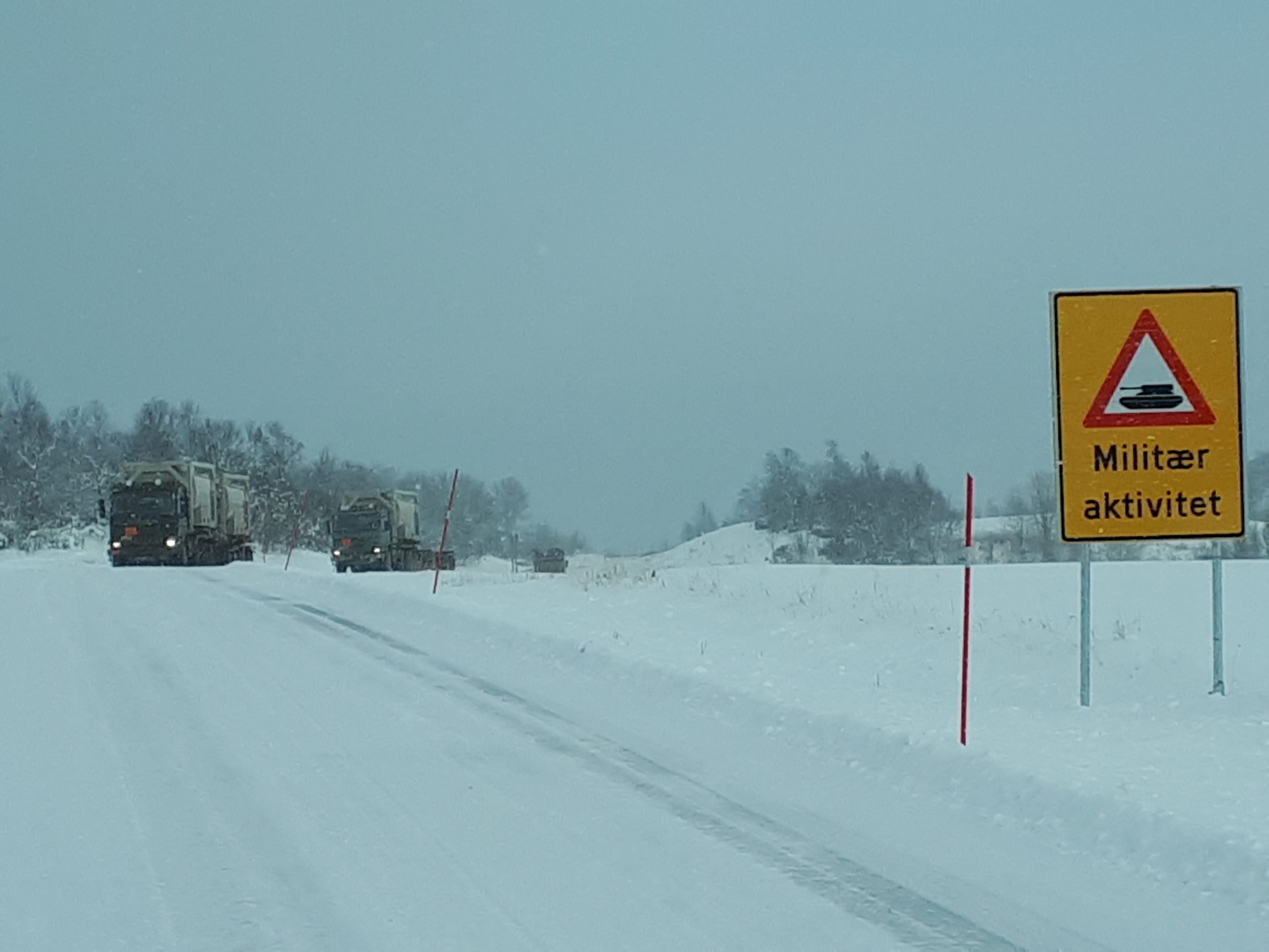Snø på bakken, et skilt hvor det står "Militær aktivitet". To militære kjøretøy sees i bakgrunnen