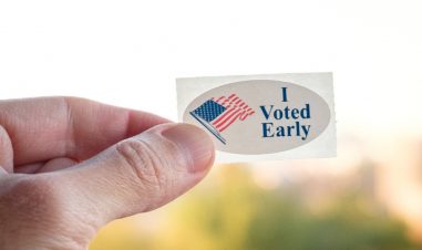 En hånd holder et klistemerke opp mot luften. På klistemerke står det "I voted early"