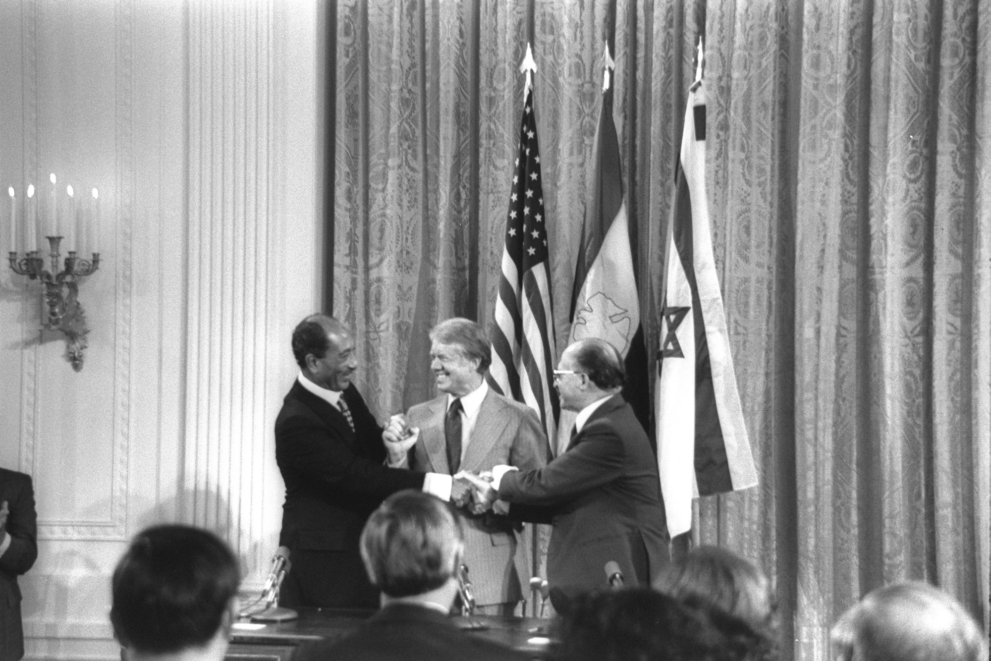 Svart-hvitt bilde av tre dresskledde menn som tar hverandre smilende i hendene. I bakgrunnen kan vi antyde både det amerikanske og israelske flagget i tillegg til et flagg til.