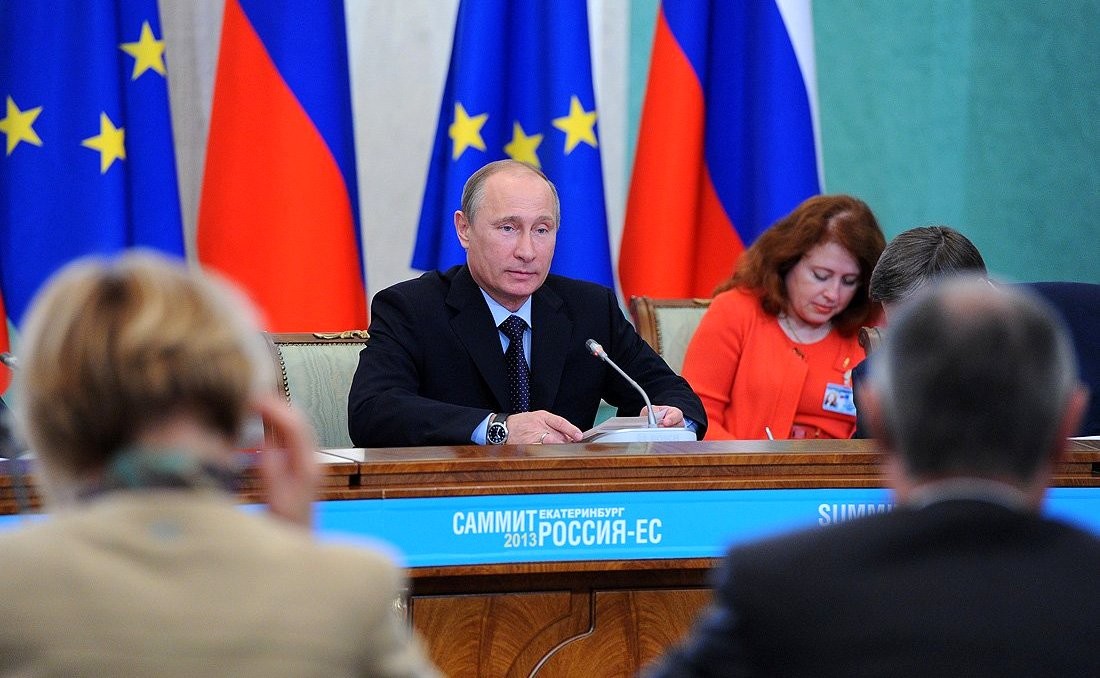 Putin sitter ved et bord. I bakgrunnen er russiske flagg og EU-flagg.