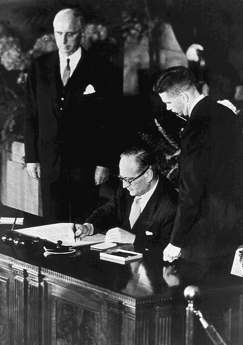 Tre menn står bak et skrivebord og signerer dokumenter