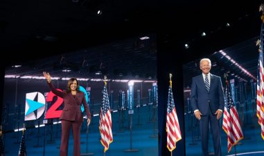 Joe Biden og Kamela Harris på en scene. Harris vinker. Biden smiler. Bak dem er det amerikanske flagg