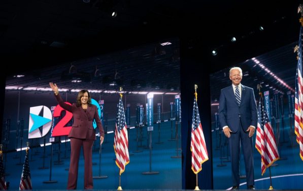 Joe Biden og Kamela Harris på en scene. Harris vinker. Biden smiler. Bak dem er det amerikanske flagg