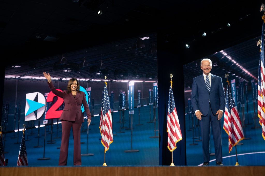 Joe Biden og Kamela Harris  på en scene. Harris vinker. Biden smiler. Bak dem er det amerikanske flagg