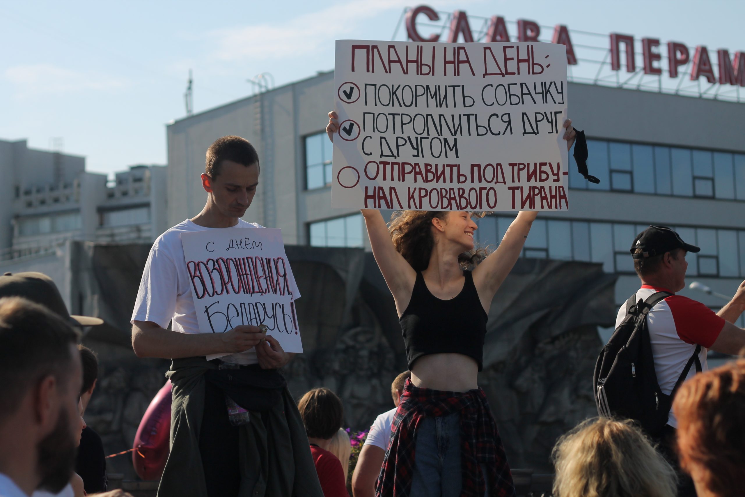 En ung mann og kvinne står med hvert sitt skilt under en demonstrasjon. Mannen står med bøyd nakke og skiltet ved brystet, kvinnen smiler og hever skiltet over hodet med begge hender.