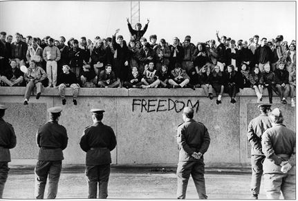 En jublende menneskemengde har klatret opp på toppen av Berlinmuren. Sikkerhetsstyrker ser på fra den andre siden.