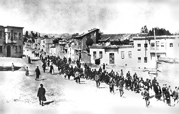 svart hvitt bilde av en rekke mennesker som går gjennom en by eskortert av vakter