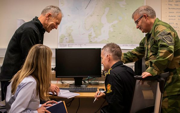 Fire personer i samtale, hvorav en militær og en politi, med ryggen delvis mot kamera.