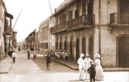 Mennesker i klær fra starten av 1900-tallet står og prater sammen i en gate dominert av flotte fasader.står og prat