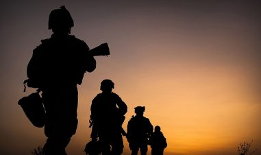 Bilde av fire soldater marsjerende på rekke vekk fra kamera. Bare omrisset er synlig i solnedgangen.