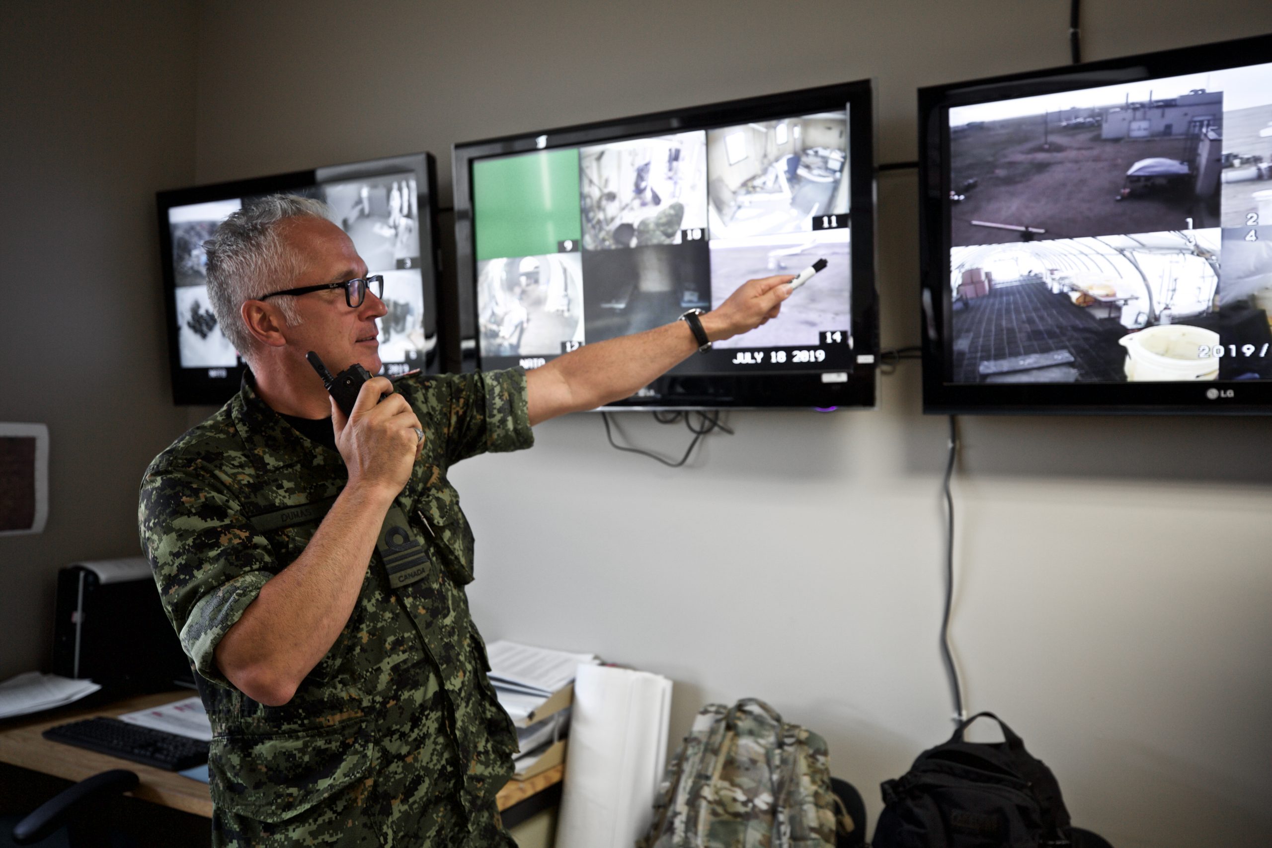 En eldre mann i militæruniform står foran tre TV-skjermer og overvåker treningsaktiviteten.