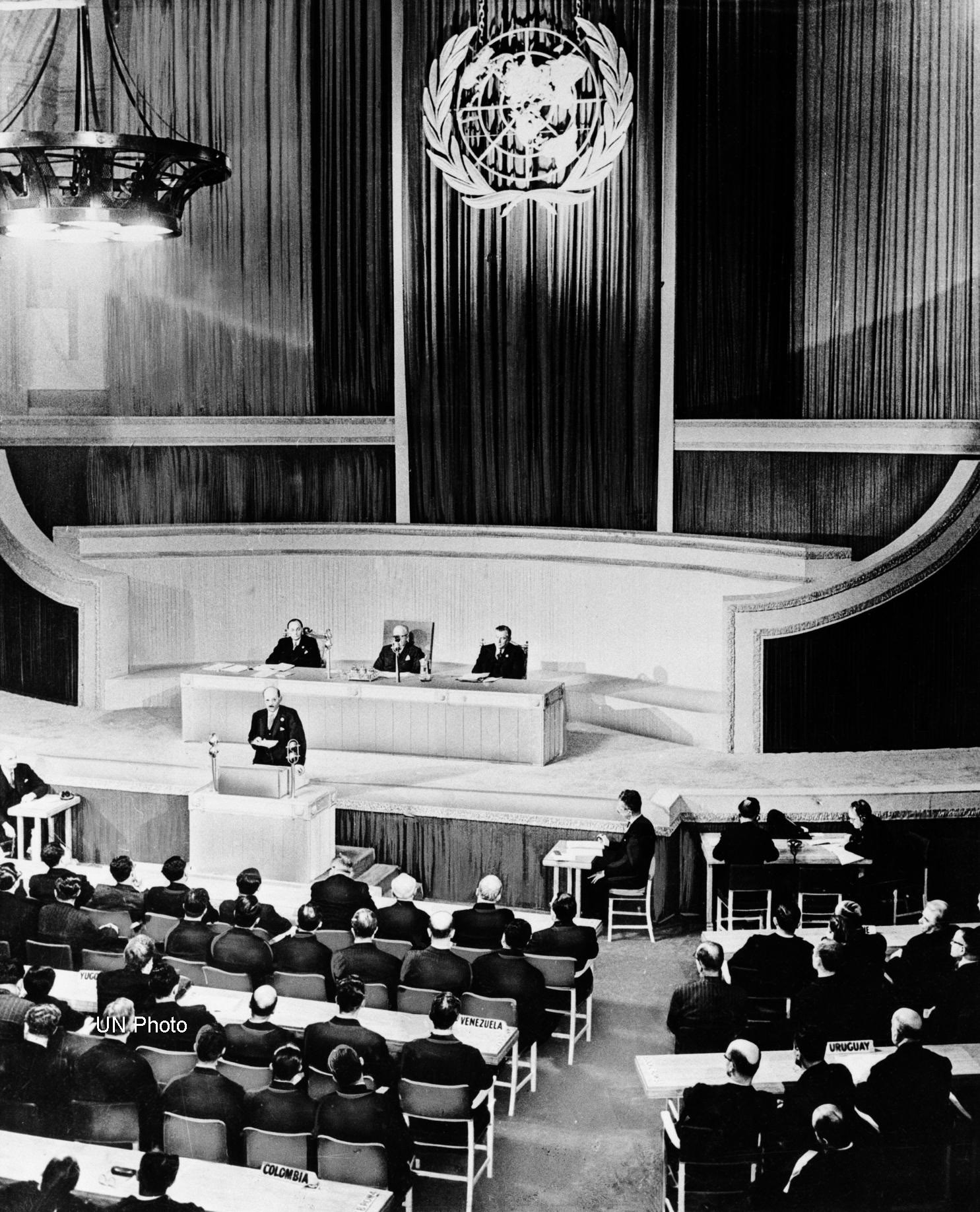 Tre menn sitter på et podie, foran dem snakker en man fra en talerstol. Bak dem henger det en enormt flagg med FNs emblem. Han taler til en sal med mange mennesker sittende på stoler.